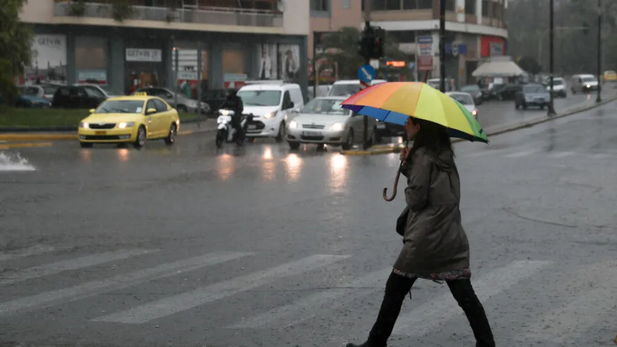 Γυναίκα με πολύχρωμη ομπρέλα διασχίζει δρόμο υπό βροχή στην Αθήνα.