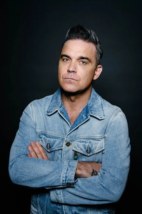 Ο Robbie Williams με τζιν τζάκετ σε σκούρο φόντο.