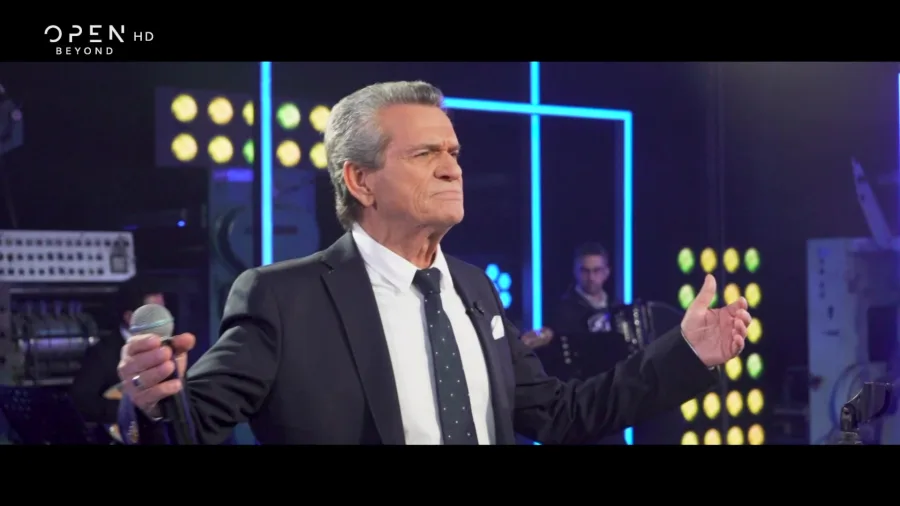Ο Γιώργος Μαργαρίτης τραγουδάει σε τηλεοπτικό πλατό κρατώντας μικρόφωνο.