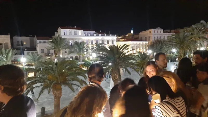 Άνθρωποι σε ταράτσα με θέα φωτισμένη πλατεία και φοίνικες τη νύχτα.