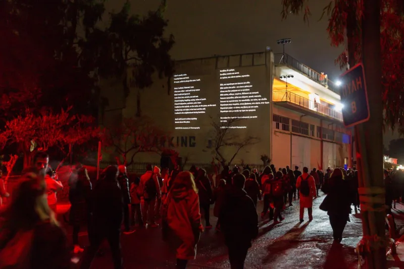Πλήθος κόσμου παρακολουθεί προβολή κειμένου σε κτίριο στην Ελευσίνα.