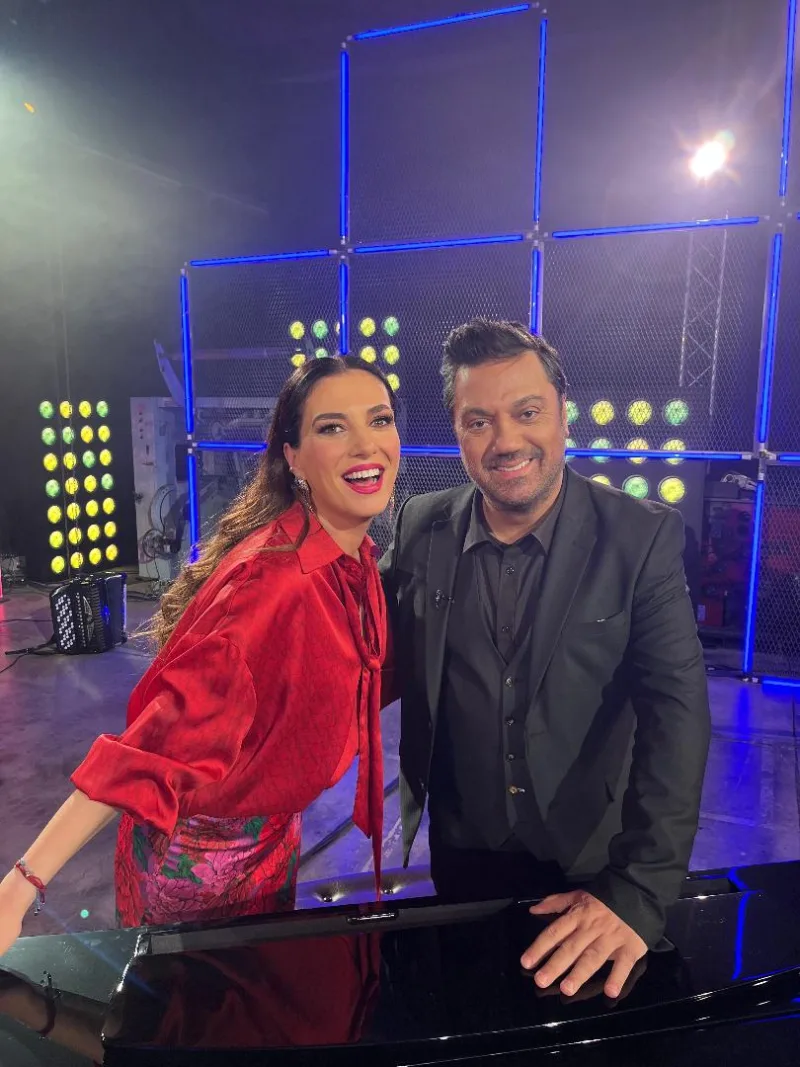 Μια γυναίκα με κόκκινο πουκάμισο και ένας άνδρας με μαύρο κοστούμι σε τηλεοπτικό πλατό.