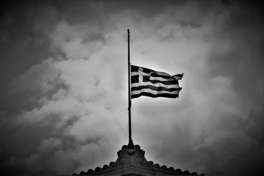 Η ελληνική σημαία κυματίζει μεσίστια σε ασπρόμαυρο φόντο με συννεφιασμένο ουρανό.