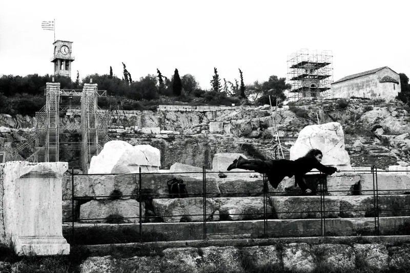 Ασπρόμαυρη φωτογραφία με άτομο να αιωρείται μπροστά από αρχαιολογικό χώρο στην Ελευσίνα.