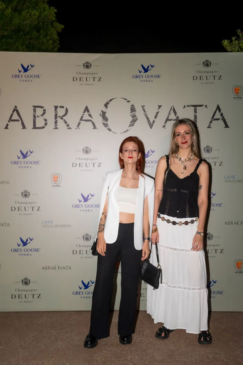 Δύο γυναίκες ποζάρουν σε opening party εστιατορίου μπροστά από διαφημιστικό τοίχο.