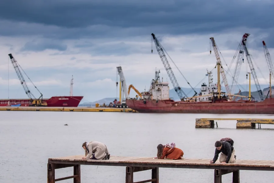 Τρία άτομα γονατιστά σε ξύλινη προβλήτα με φόντο λιμάνι και γερανούς.