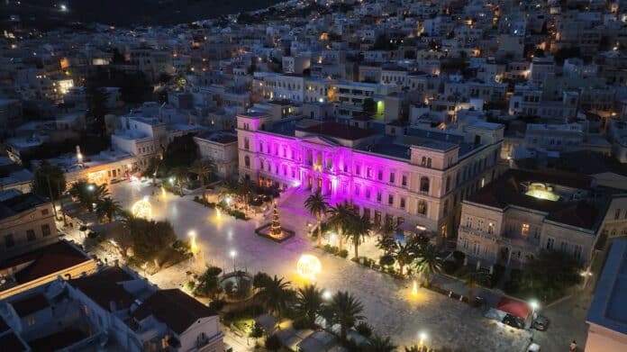 Νυχτερινή άποψη πλατείας με φωταγωγημένο κτίριο σε μωβ χρώμα.