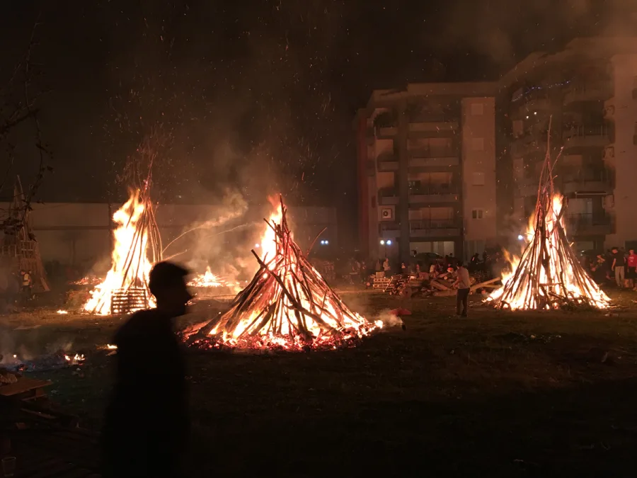 Μεγάλες φωτιές σε ανοιχτό χώρο τη νύχτα με κόσμο και πολυκατοικίες στο βάθος.