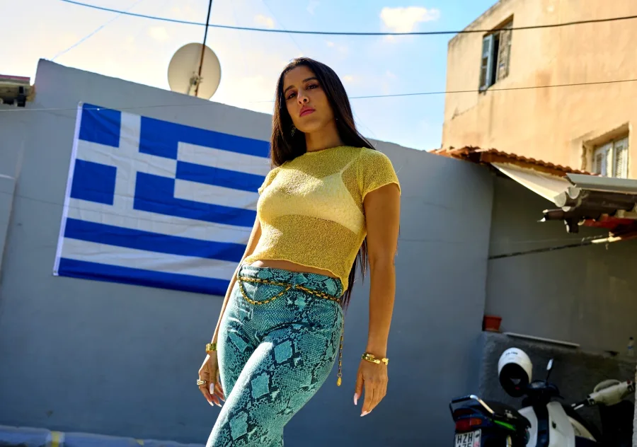 Η Μαρίνα Σάττι με κίτρινο τοπ και παντελόνι με animal print μπροστά από την ελληνική σημαία.