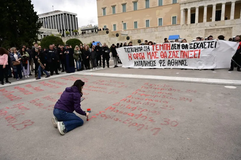 Γυναίκα γράφει με κόκκινη μπογιά ονόματα στο Σύνταγμα μπροστά από πανό για τα Τέμπη.