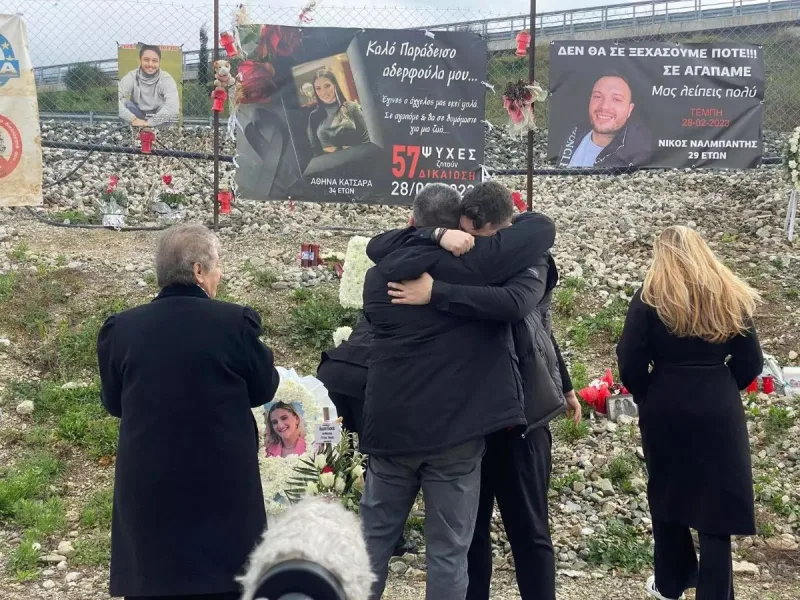Άνθρωποι αγκαλιάζονται σε μνημόσυνο στα Τέμπη μπροστά από πανό με φωτογραφίες θυμάτων.