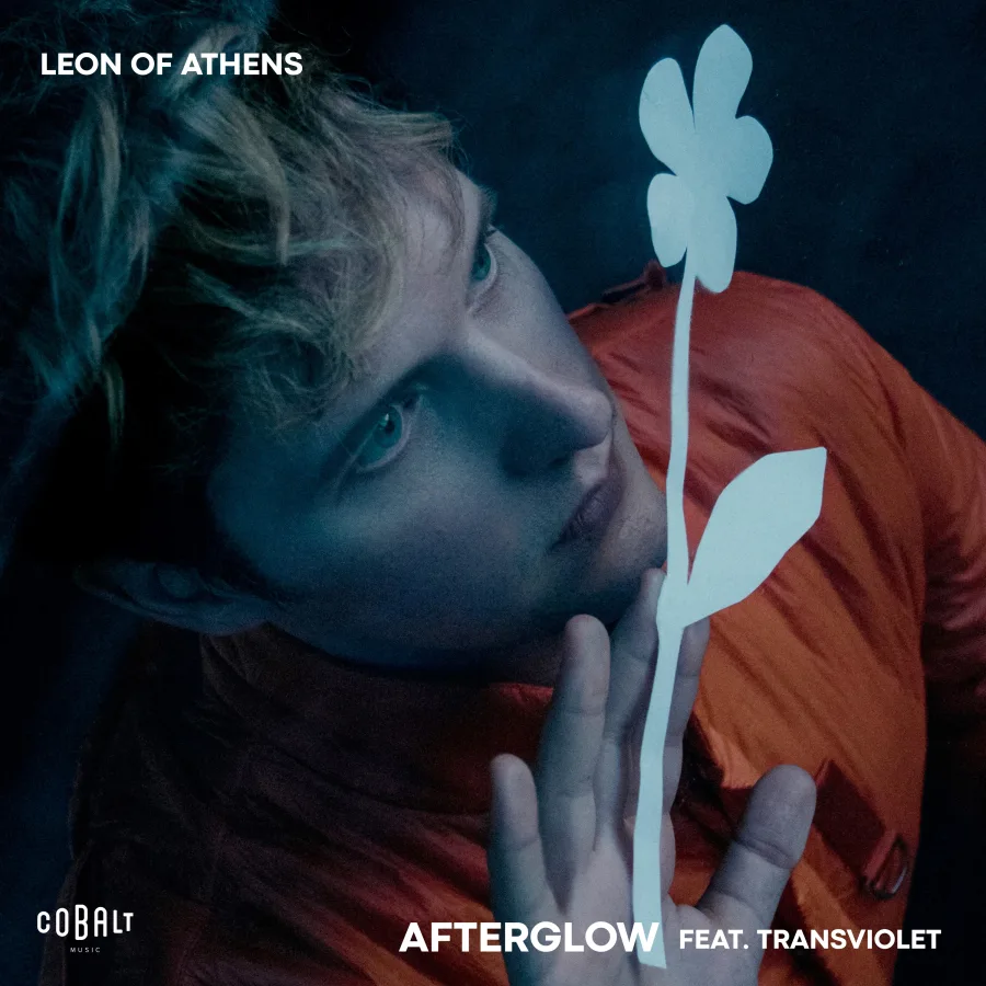 Ο Leon Of Athens σε κοντινό πλάνο με πορτοκαλί ρούχο και μια χάρτινη μαργαρίτα.