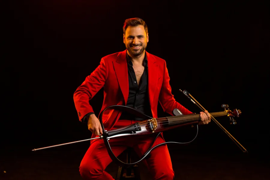 Ο HAUSER με κόκκινο κοστούμι κρατάει ένα ηλεκτρικό τσέλο.