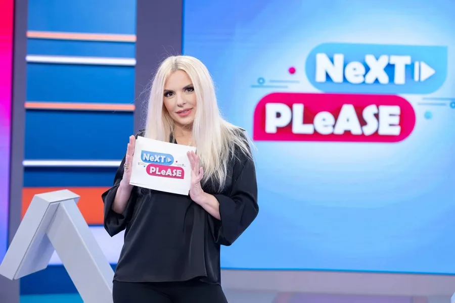 Η Αννίτα Πάνια σε τηλεοπτικό πλατό κρατώντας κάρτα με το λογότυπο Next Please.