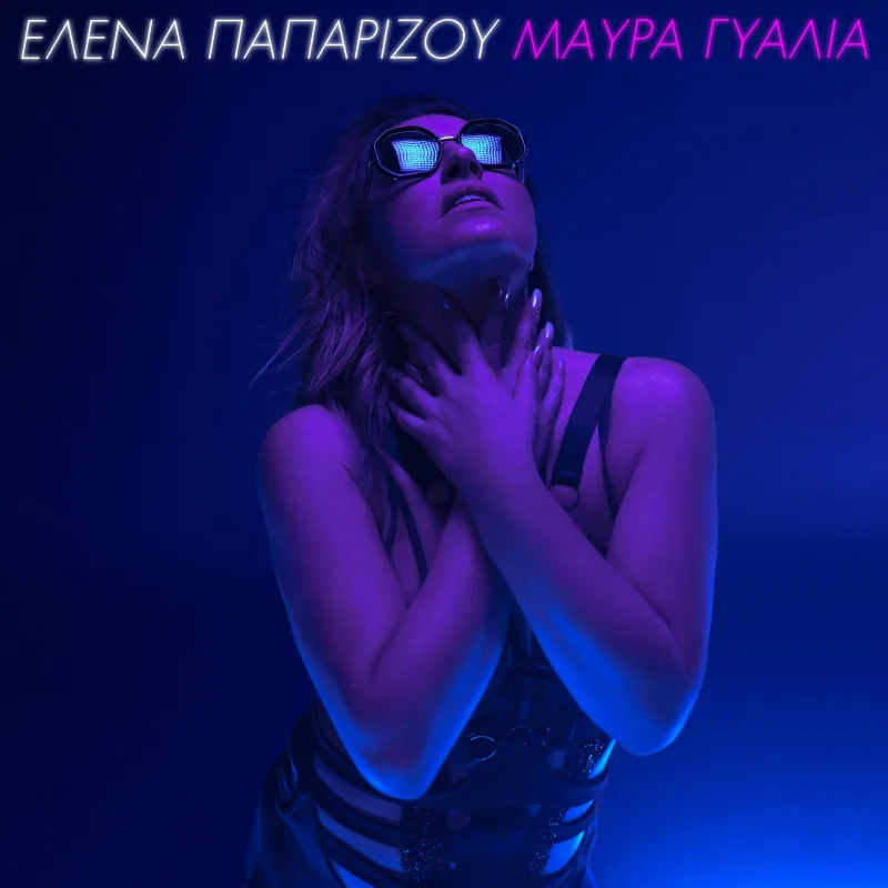 Η Έλενα Παπαρίζου με μαύρα γυαλιά και χέρια στον λαιμό σε μπλε και μωβ φωτισμό.