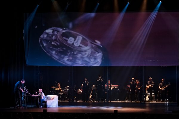 Σκηνή από την παράσταση The Wall - Pink Floyd's Rock Opera με μουσικούς και προβολή σε οθόνη.