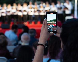 Θεατής φωτογραφίζει με το κινητό του μια χορωδία σε υπαίθρια συναυλία.