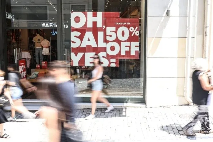 Βιτρίνα καταστήματος με κόκκινη αφίσα για θερινές εκπτώσεις 50% και περαστικούς στον δρόμο.