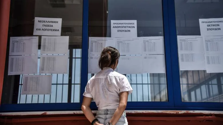Μαθήτρια κοιτάζει αναρτημένες λίστες με αποτελέσματα εξετάσεων σε τζαμαρία σχολείου.