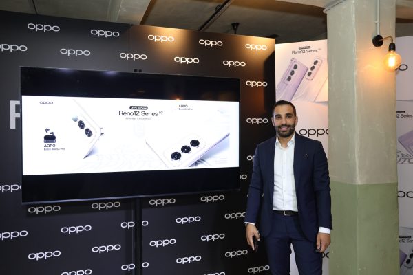 Άνδρας με κοστούμι δίπλα σε οθόνη παρουσίασης της σειράς OPPO Reno12.