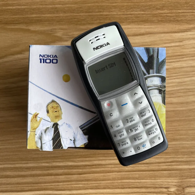 Κινητό τηλέφωνο Nokia 1100 πάνω στο κουτί του σε ξύλινη επιφάνεια.