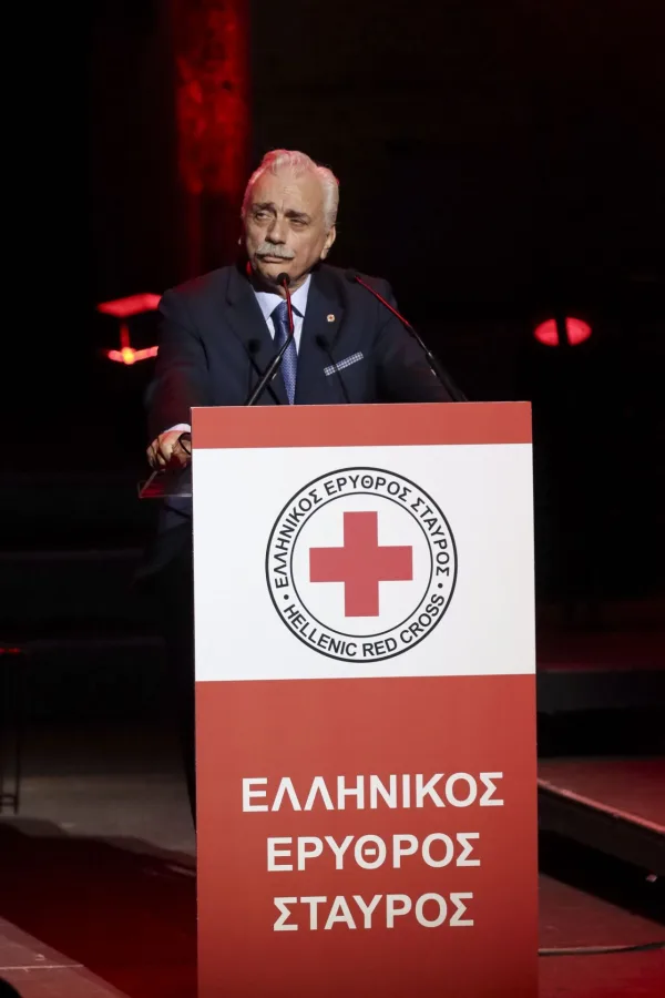 Άνδρας με κοστούμι μιλάει σε πόντιουμ του Ελληνικού Ερυθρού Σταυρού.