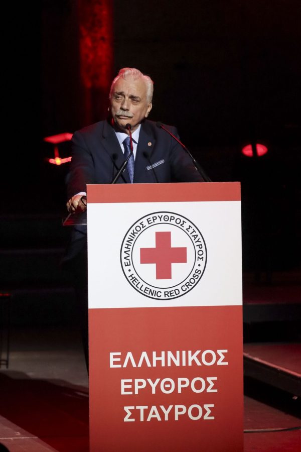 Άνδρας με κοστούμι μιλάει σε πόντιουμ του Ελληνικού Ερυθρού Σταυρού.