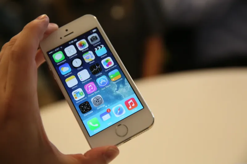 Ένα λευκό iPhone 5s στο χέρι ενός χρήστη με την αρχική οθόνη του iOS 7.