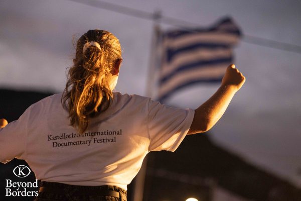Κοπέλα με μπλουζάκι του Kastellorizo International Documentary Festival μπροστά από την ελληνική σημαία.