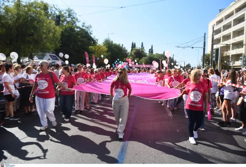 Πλήθος κόσμου με ροζ μπλουζάκια κρατά μια μεγάλη ροζ κορδέλα στον δρόμο για το Greece Race for the Cure.