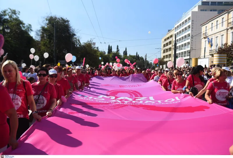 Πλήθος κόσμου με ροζ μπλούζες κρατά μια μεγάλη ροζ σημαία στο Greece Race for the Cure.