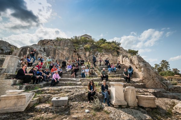 Ομάδα ανθρώπων κάθεται στα πέτρινα σκαλιά ενός αρχαιολογικού χώρου στην Ελευσίνα.