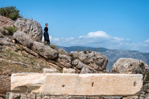 Καλλιτέχνης με μαύρα ρούχα στέκεται σε βραχώδη πλαγιά αρχαιολογικού χώρου.