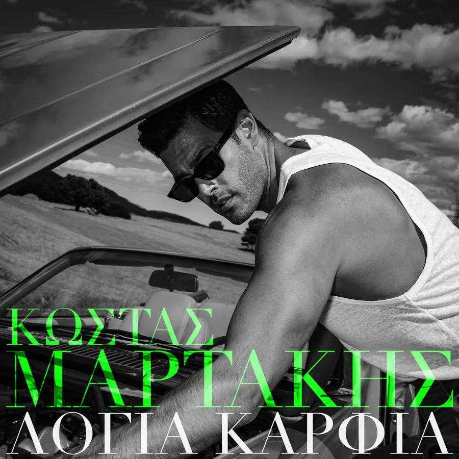 Ο Κώστας Μαρτάκης με γυαλιά ηλίου και λευκό φανελάκι δίπλα σε αυτοκίνητο.