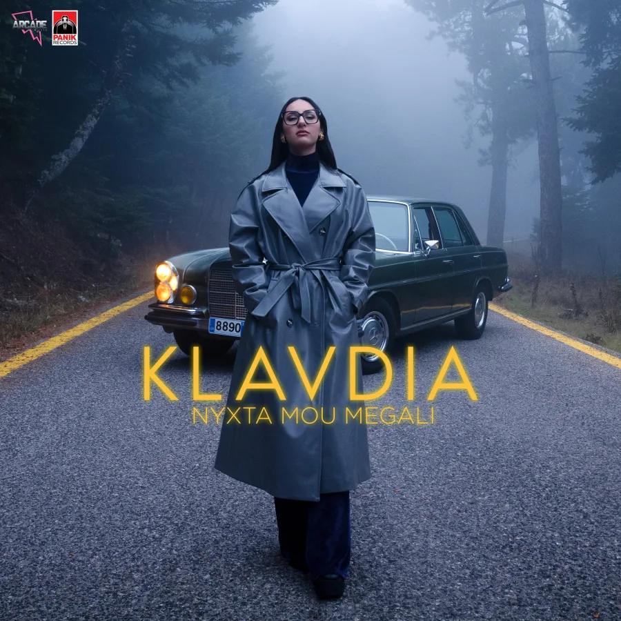 Η Klavdia με γκρι καμπαρντίνα μπροστά από κλασικό αυτοκίνητο σε ομιχλώδη δρόμο.
