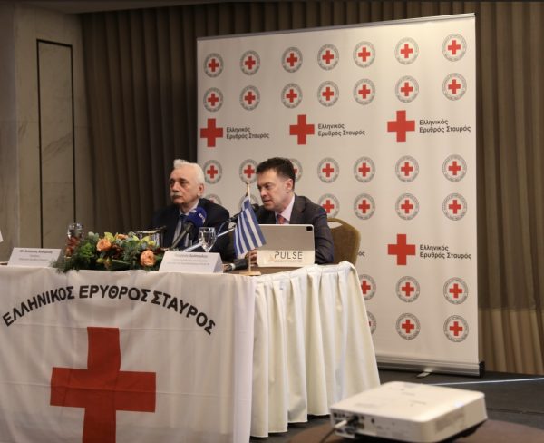 Δύο άνδρες σε συνέντευξη τύπου του Ελληνικού Ερυθρού Σταυρού με φόντο το λογότυπο του οργανισμού.