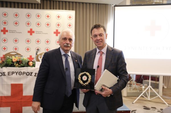 Δύο άνδρες σε εκδήλωση του Ελληνικού Ερυθρού Σταυρού κρατούν τιμητική πλακέτα.