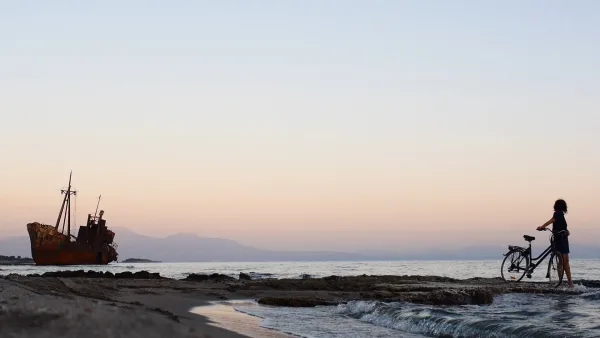 Μια γυναίκα με ποδήλατο σε παραλία δίπλα στη θάλασσα, με ένα σκουριασμένο ναυάγιο πλοίου στο βάθος κατά το ηλιοβασίλεμα.