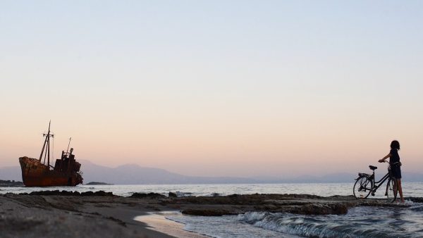 Μια γυναίκα με ποδήλατο σε παραλία δίπλα στη θάλασσα, με ένα σκουριασμένο ναυάγιο πλοίου στο βάθος κατά το ηλιοβασίλεμα.