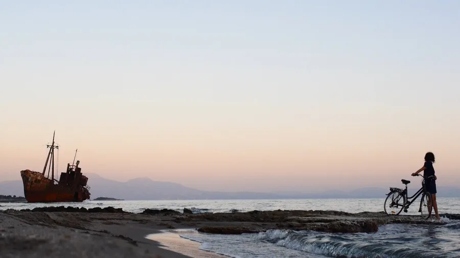 Μια γυναίκα με ποδήλατο στην ακτή με φόντο ένα σκουριασμένο ναυάγιο πλοίου κατά το ηλιοβασίλεμα.