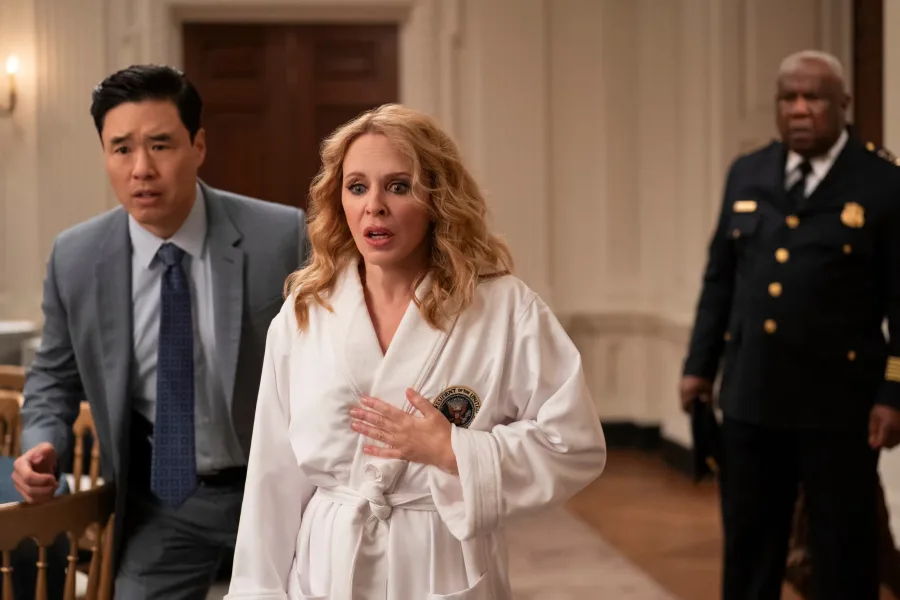 Η Kylie Minogue με λευκό μπουρνούζι και έκφραση έκπληξης δίπλα σε δύο άνδρες.