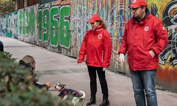 Εθελοντές του Ελληνικού Ερυθρού Σταυρού προσεγγίζουν άστεγο με σκύλο σε δρόμο με γκράφιτι.
