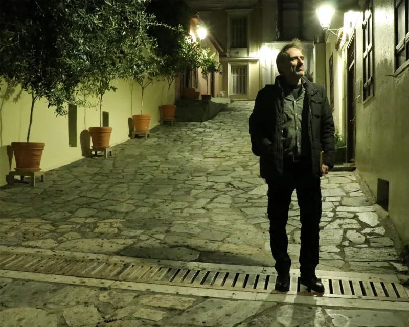 Άνδρας με μαύρο μπουφάν στέκεται σε πλακόστρωτο δρομάκι τη νύχτα.