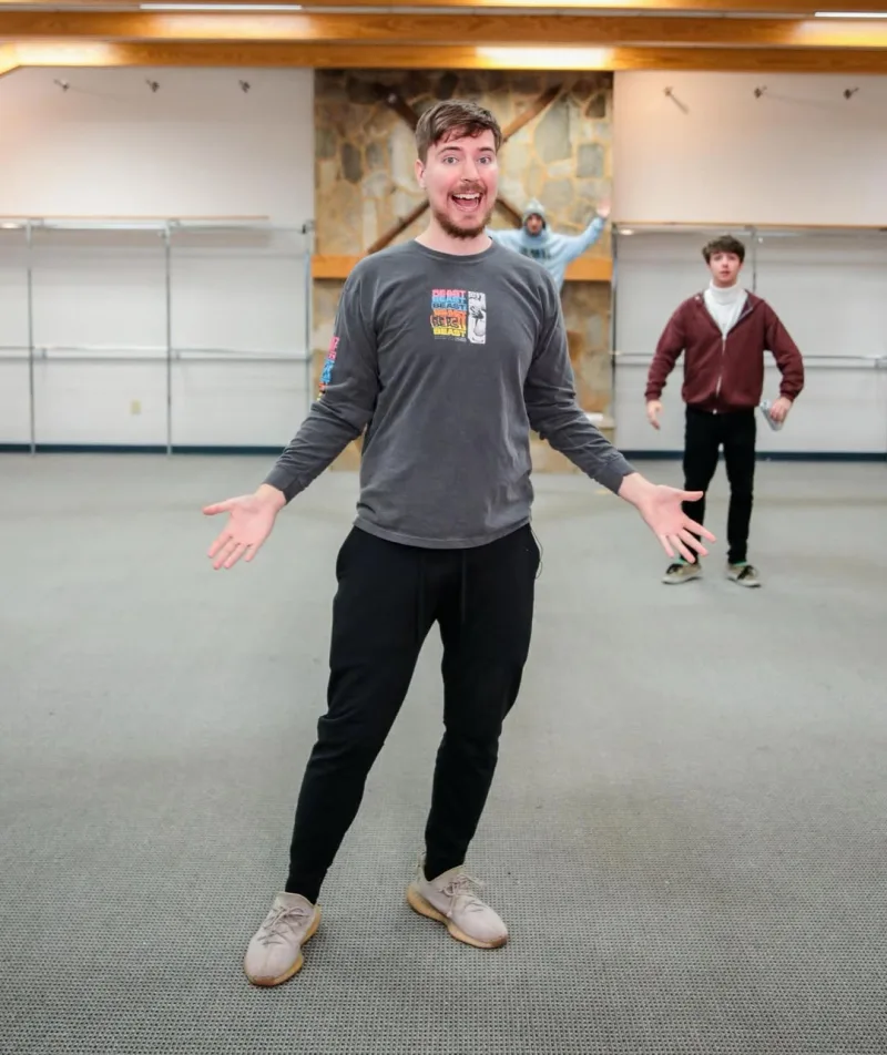 Ο MrBeast με ανοιχτά χέρια σε έναν μεγάλο άδειο χώρο, φορώντας γκρι μπλούζα και μαύρο παντελόνι.