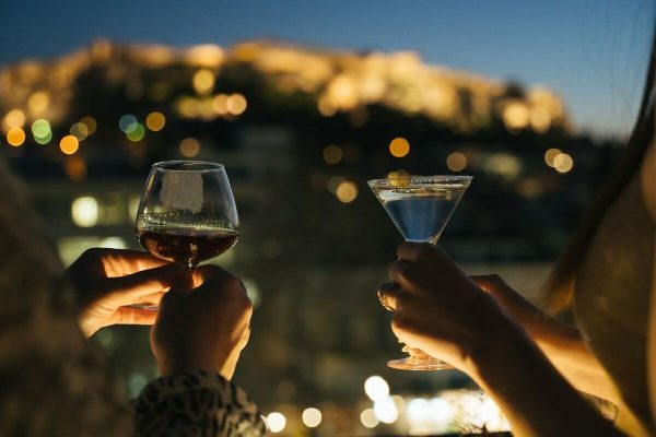 Δύο χέρια κρατούν ποτήρια με κοκτέιλ και κρασί σε rooftop bar της Αθήνας με θέα την Ακρόπολη.