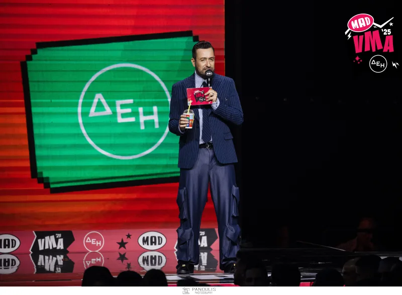 Ο παρουσιαστής των Mad Video Music Awards 2025 από την ΔΕΗ κρατάει μικρόφωνο και ποτήρι στην σκηνή.
