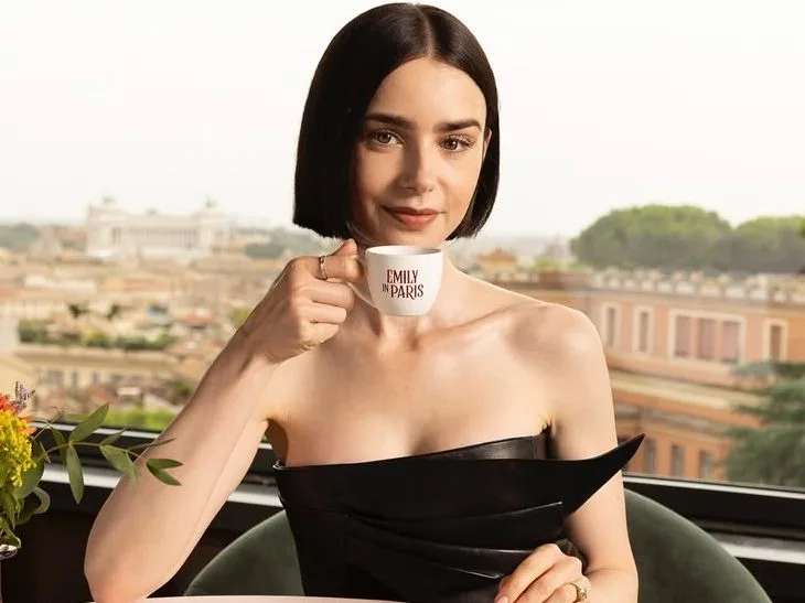 Η Lily Collins κρατά φλιτζάνι καφέ με το λογότυπο "Emily in Paris", απαντώντας αινιγματικά στην ερώτηση για την παρουσία της στην Ελλάδα.