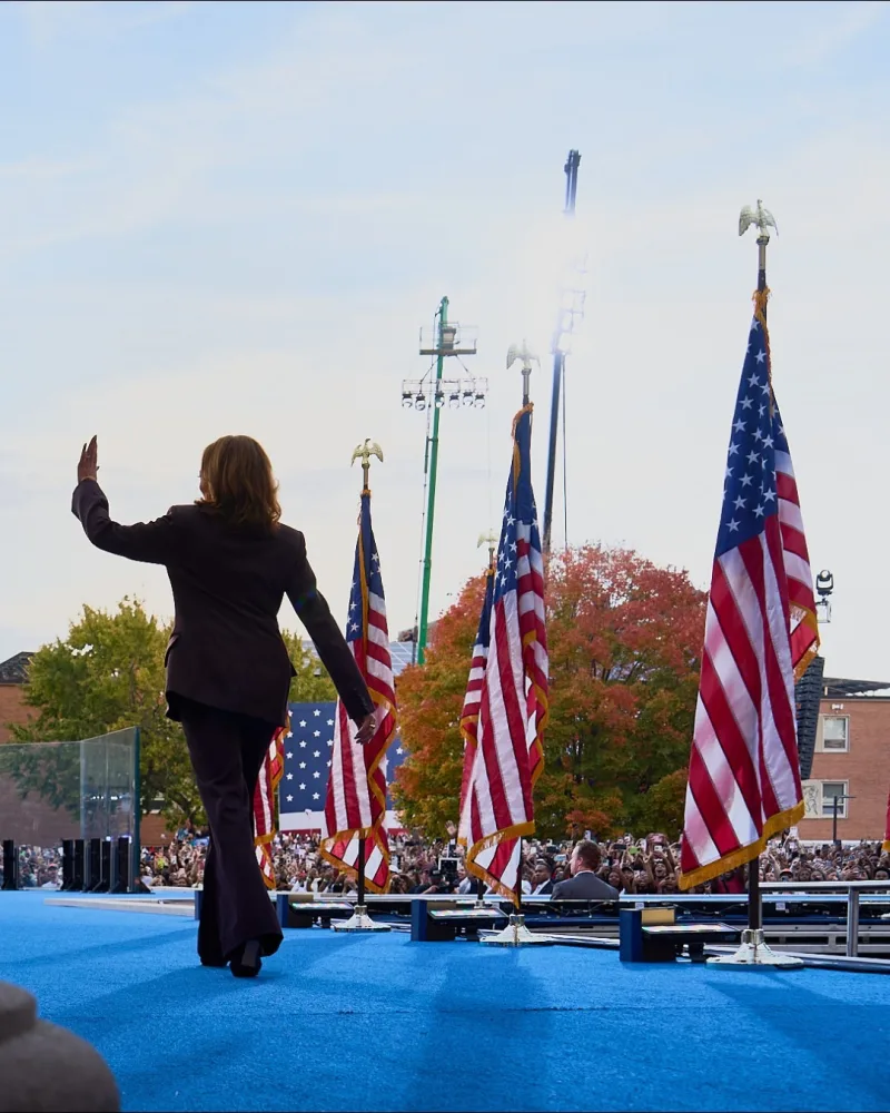 Η Kamala Harris χαιρετά το πλήθος σε προεκλογική συγκέντρωση με αμερικανικές σημαίες.
