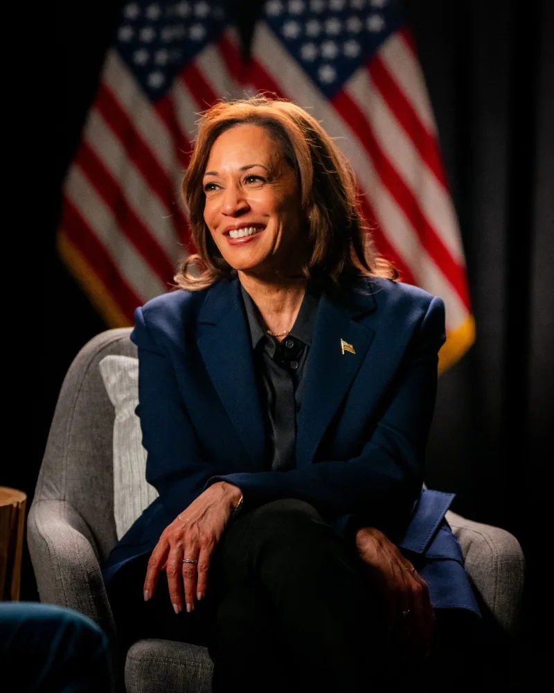 Η Kamala Harris χαμογελαστή σε συνέντευξη με φόντο αμερικανικές σημαίες.