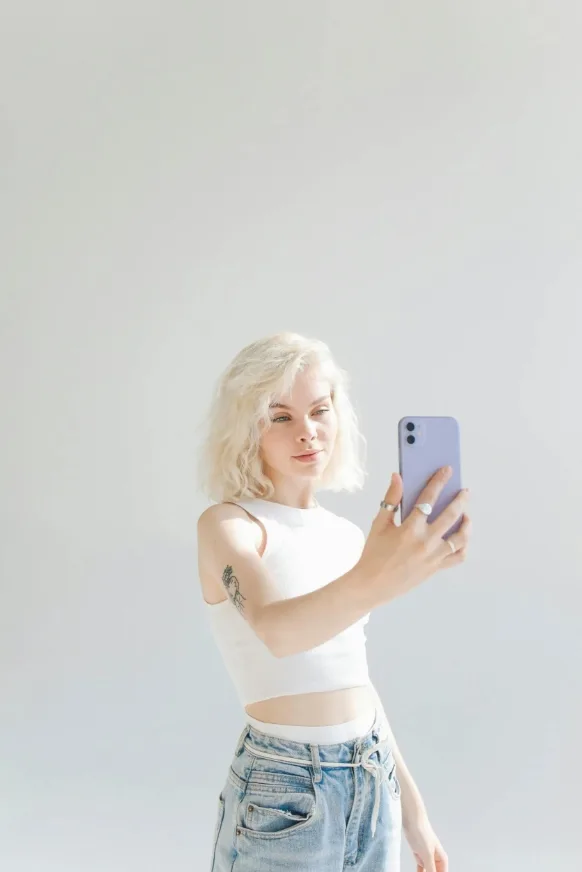Ξανθιά κοπέλα βγάζει selfie με το κινητό της σε λευκό φόντο.